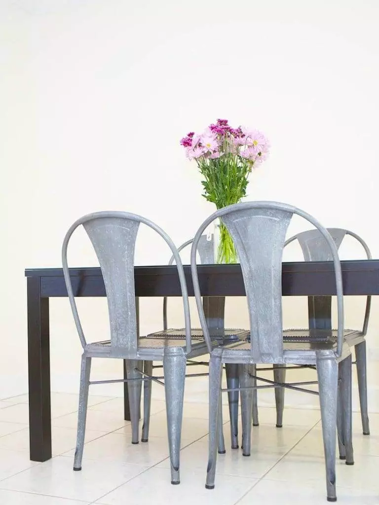 A modern dining room setup with a dark wooden table and three metal chairs evokes the elegance of Martinique. A vase with pink and purple flowers graces the table, while the background wall in a light cream color provides a serene backdrop.
