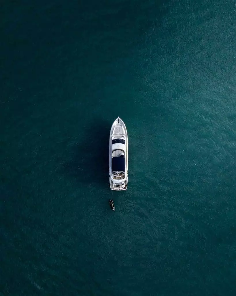 Aerial view of a yacht anchored in serene, deep blue ocean waters. A small boat is positioned alongside the yacht, creating a tranquil scene against the vast expanse of water.