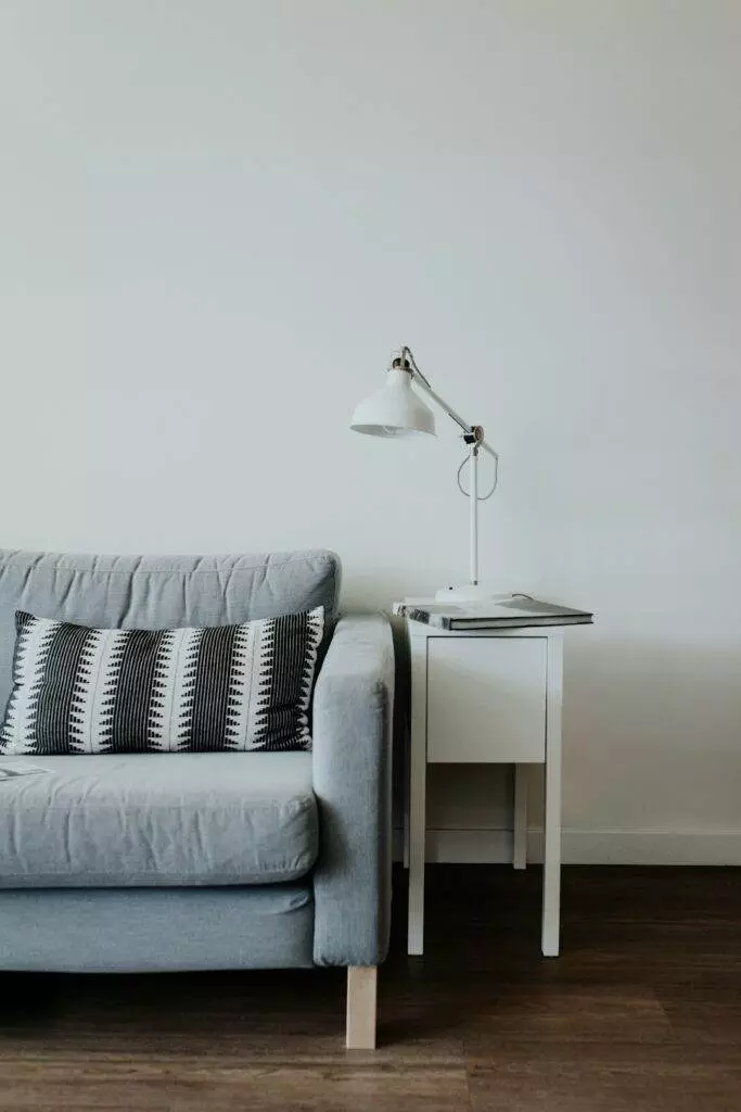 A minimalist living room features a light gray sofa with patterned cushions. Next to it is a white side table holding a white desk lamp and closed book, set against a plain white wall and wooden floor.