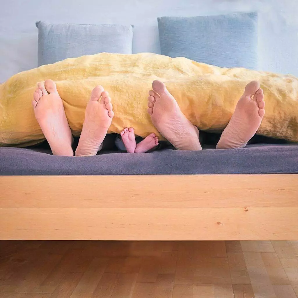 Three pairs of feet—two adults and a child—peek out from under a yellow blanket on a wooden bed, suggesting a family lying together.