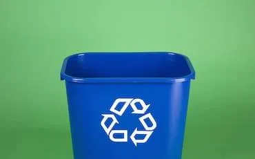 A blue recycling bin with a white recycling symbol on the front, set against a plain green background.
