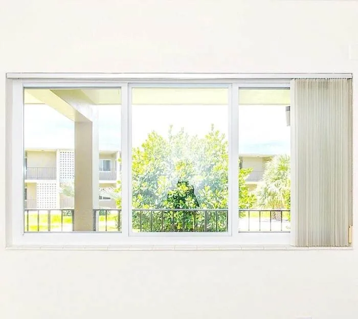 View through a large window with white frames, showing green trees and parts of a building outside. Vertical blinds are partially open on the right side. Bright, sunny day with a clear sky.