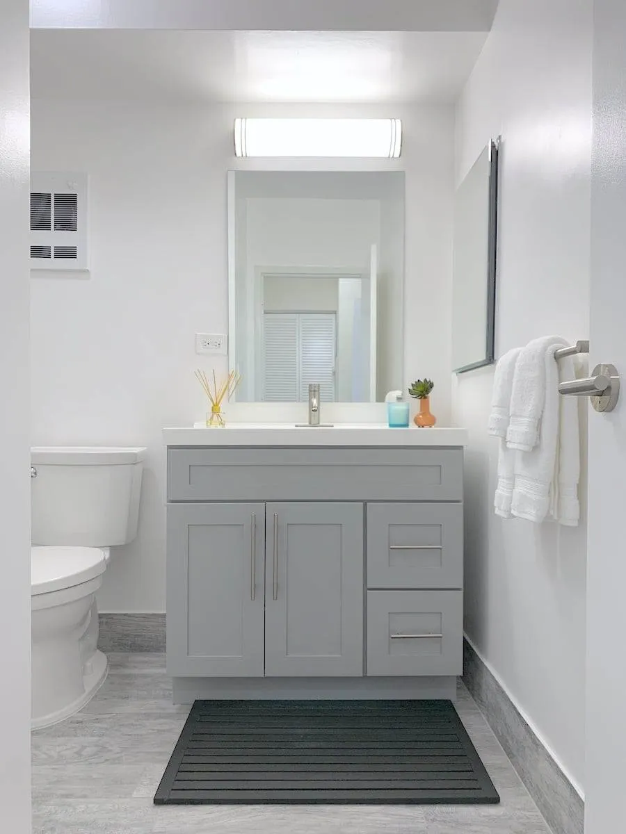 A modern, bright apartment bathroom features a gray vanity with a mirror above. It boasts a white countertop and sink, adorned with folded towels. A toilet sits to the left, while a small plant and diffuser add charm to the space.