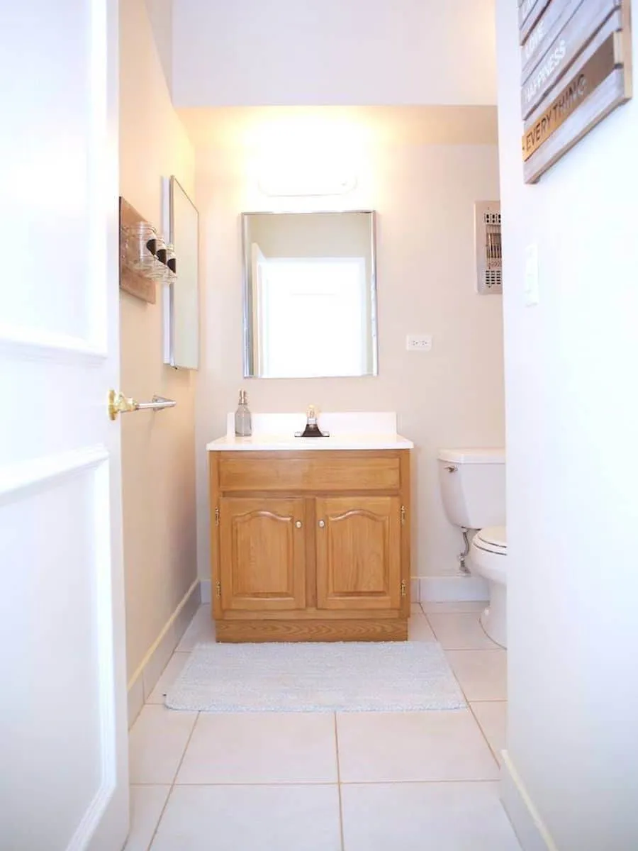 A small bathroom with light beige walls, reminiscent of a serene retreat in Martinique. It features a wooden vanity with a white sink, a rectangular mirror above, and a toilet to the right. Bright overhead lighting and a woven mat add warmth in front of the vanity.