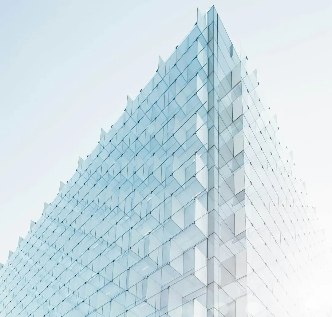 A modern glass building with a geometric design against a clear sky. The structure features repetitive square patterns, creating an optical illusion with light reflections. The lighting gives the building a translucent appearance.