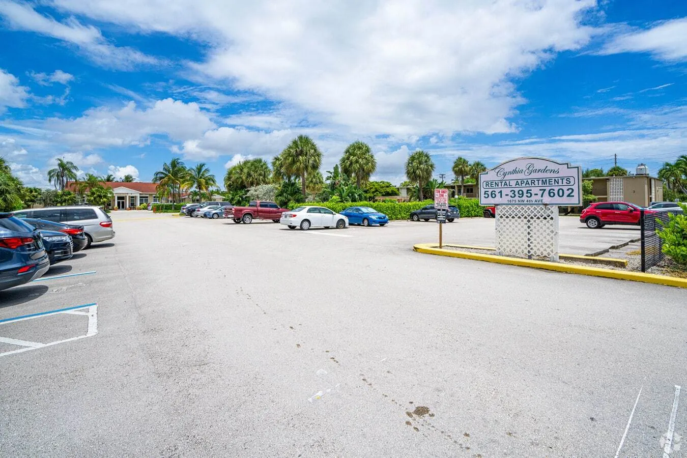 A parking lot with several cars sits under a partly cloudy sky. To the right, a sign reads 