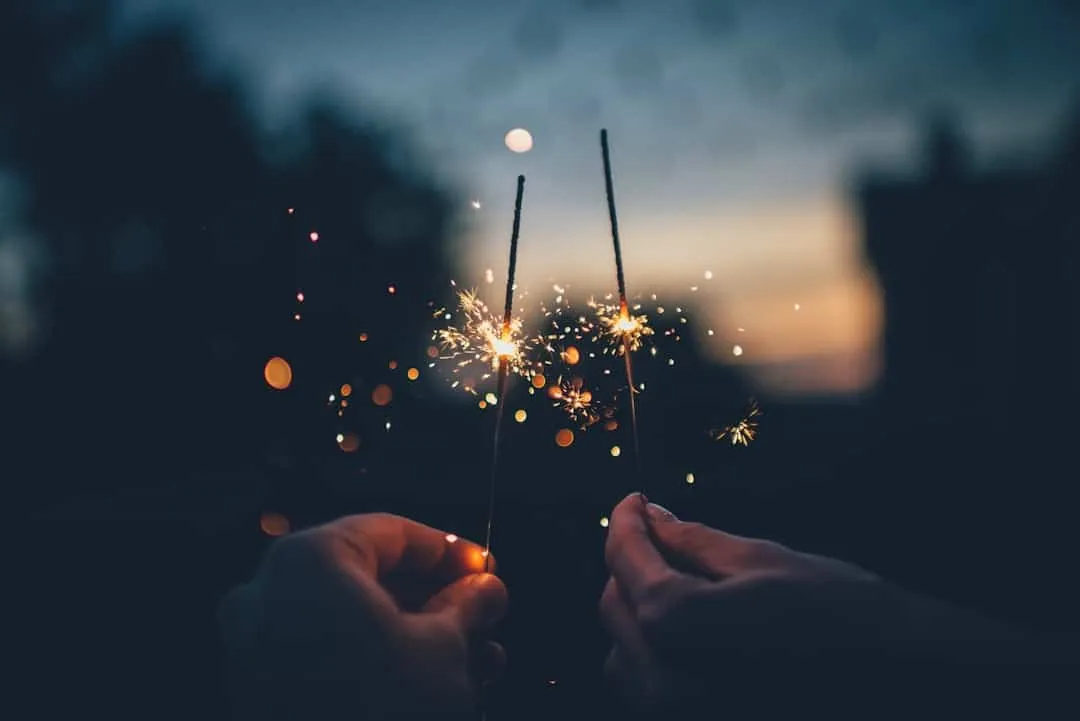 Two hands holding sparklers illuminate the evening sky. The sparklers create a bright, sparkling effect against a dark, blurred background, producing a festive and lively atmosphere.