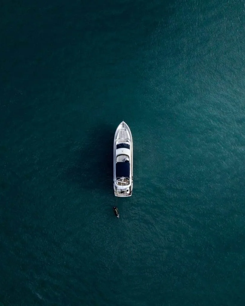Aerial view of a white yacht anchored in deep blue-green water. A small dinghy is tethered to the side of the yacht, creating a tranquil scene in the open sea.