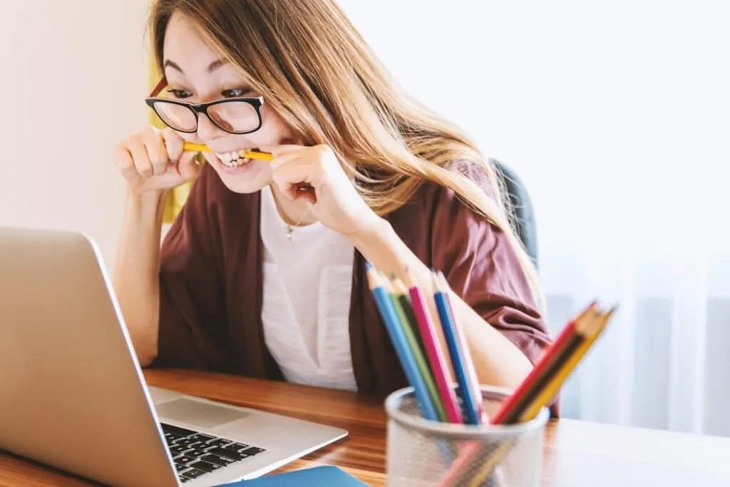 A woman with long hair and glasses bites a pencil while looking at a laptop screen. She is seated at a wooden desk with a container of colored pencils and a notebook. The room is lit by natural light from a window.
