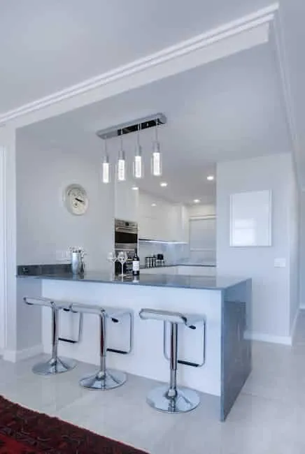 Sleek modern kitchen with a gray countertop and three chrome bar stools. A hanging light fixture with three pendant lights is above the bar. An oven is built into the cabinets, and a clock hangs on the wall. The floor is white with a red rug.