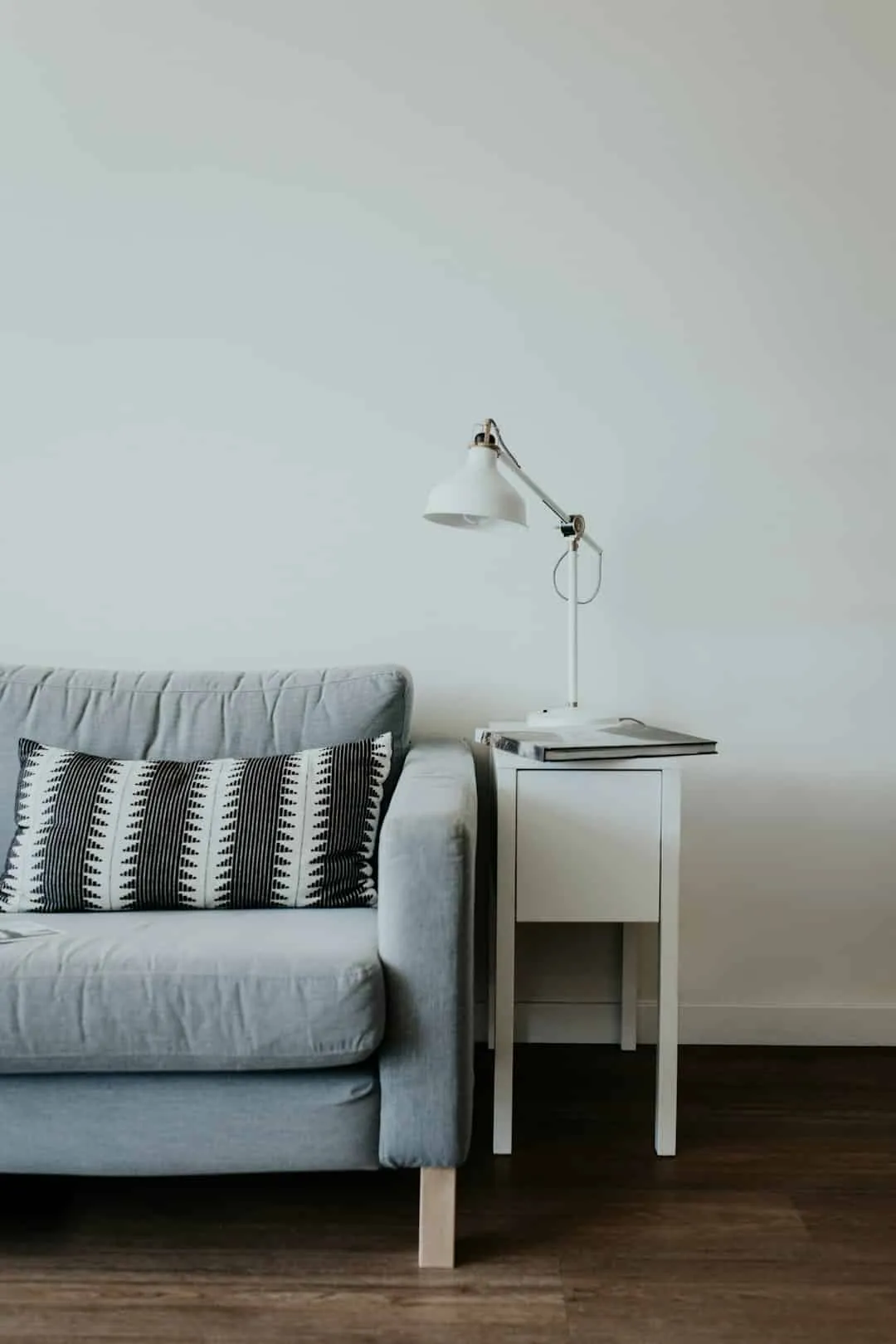 A minimalist living room features a light gray sofa with patterned cushions. Next to it is a white side table holding a white desk lamp and closed book, set against a plain white wall and wooden floor.