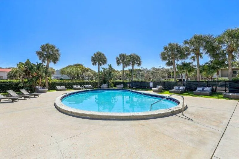 A tranquil outdoor swimming pool at Boca Raton apartments for rent, surrounded by lounge chairs and palm trees under a clear blue sky. The concrete deck is perfectly empty, creating a serene and inviting atmosphere.