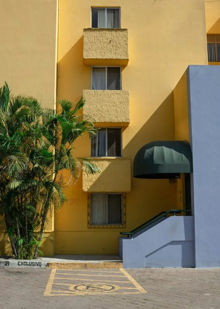 A yellow building with three square windows stands beside a palm tree. A green awning shades a ramp entrance, and a nearby parking space marked “Exclusivo” has an accessibility symbol painted on the ground.