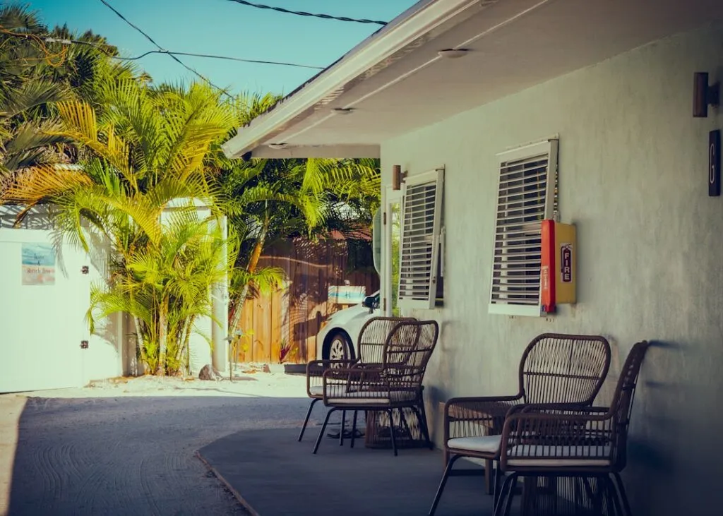 Outdoor seating area with wicker chairs lined up outside a light-colored building, with palm trees and a car partially visible in the background on a sunny day.