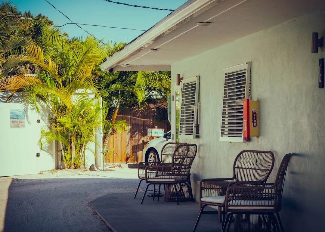 Outdoor seating area with wicker chairs lined up outside a light-colored building, with palm trees and a car partially visible in the background on a sunny day.