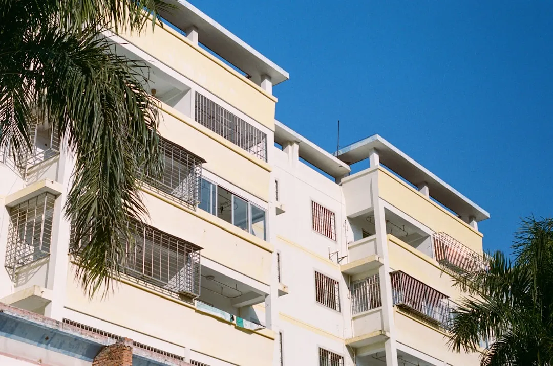 A pale yellow and white apartment building with balconies and metal railings, set against a clear blue sky. Palm tree branches are visible in the foreground.