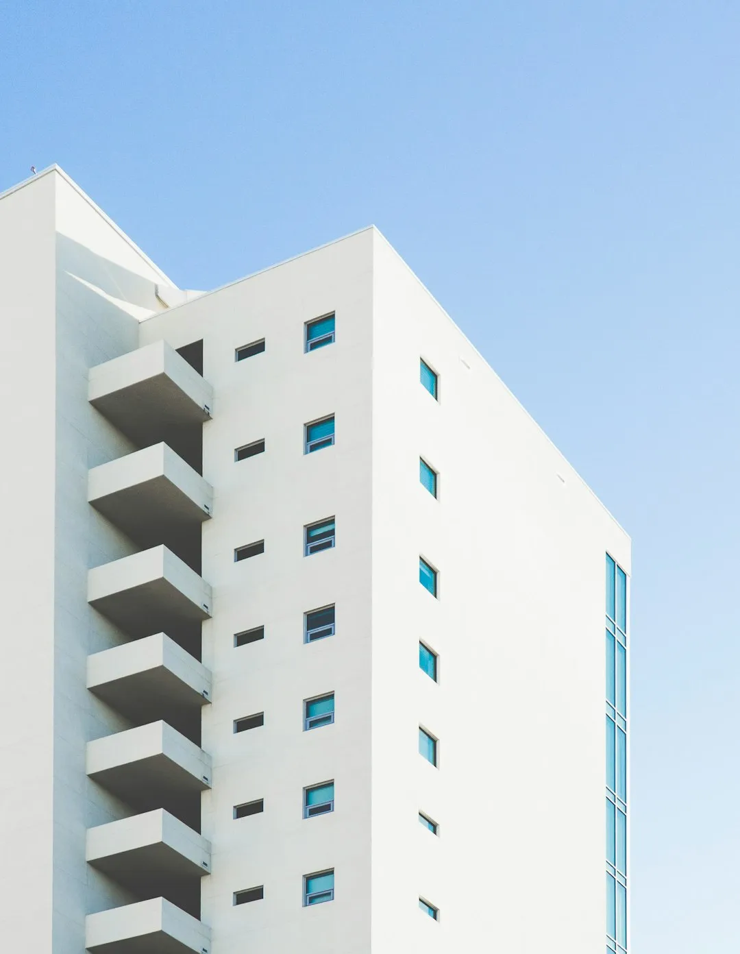 A modern white building with small square windows and protruding balconies, set against a clear blue sky. The minimalist architecture features clean lines and a smooth facade.