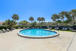 A round outdoor swimming pool, surrounded by lounge chairs and palm trees under a clear blue sky, offers relaxing seating areas and lush greenery—the perfect escape at these Boca Raton apartments.